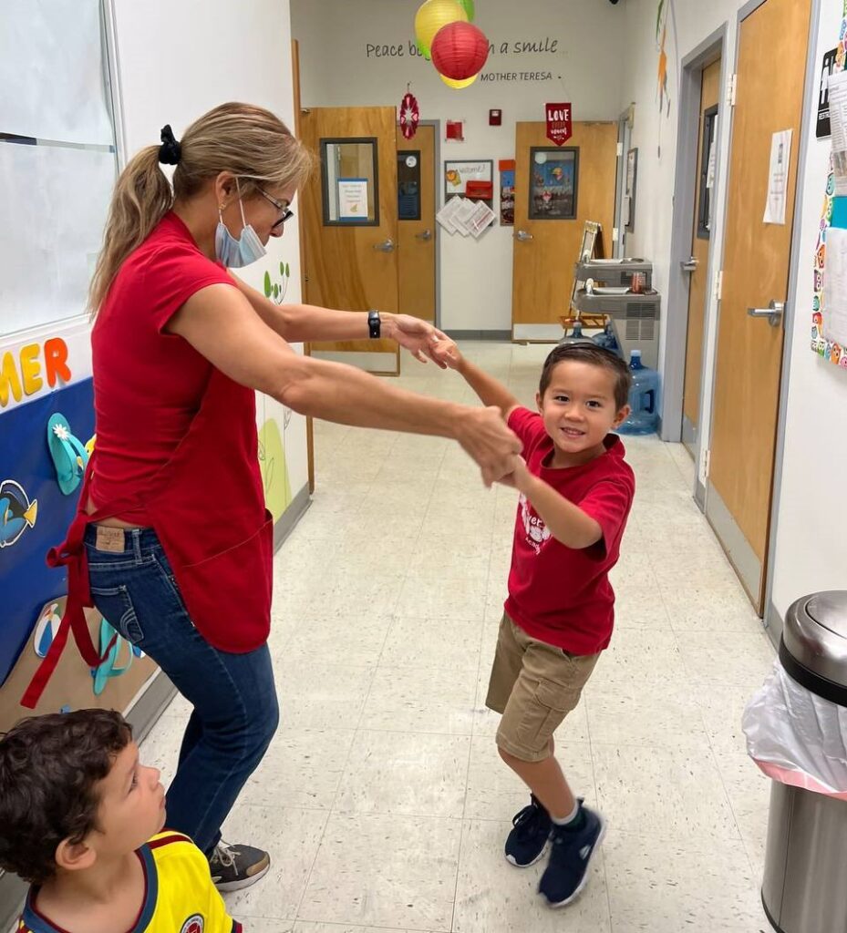 Preschool teacher dancing with toddler wearing a red t-shirt from school, in a corridor of the pre school.