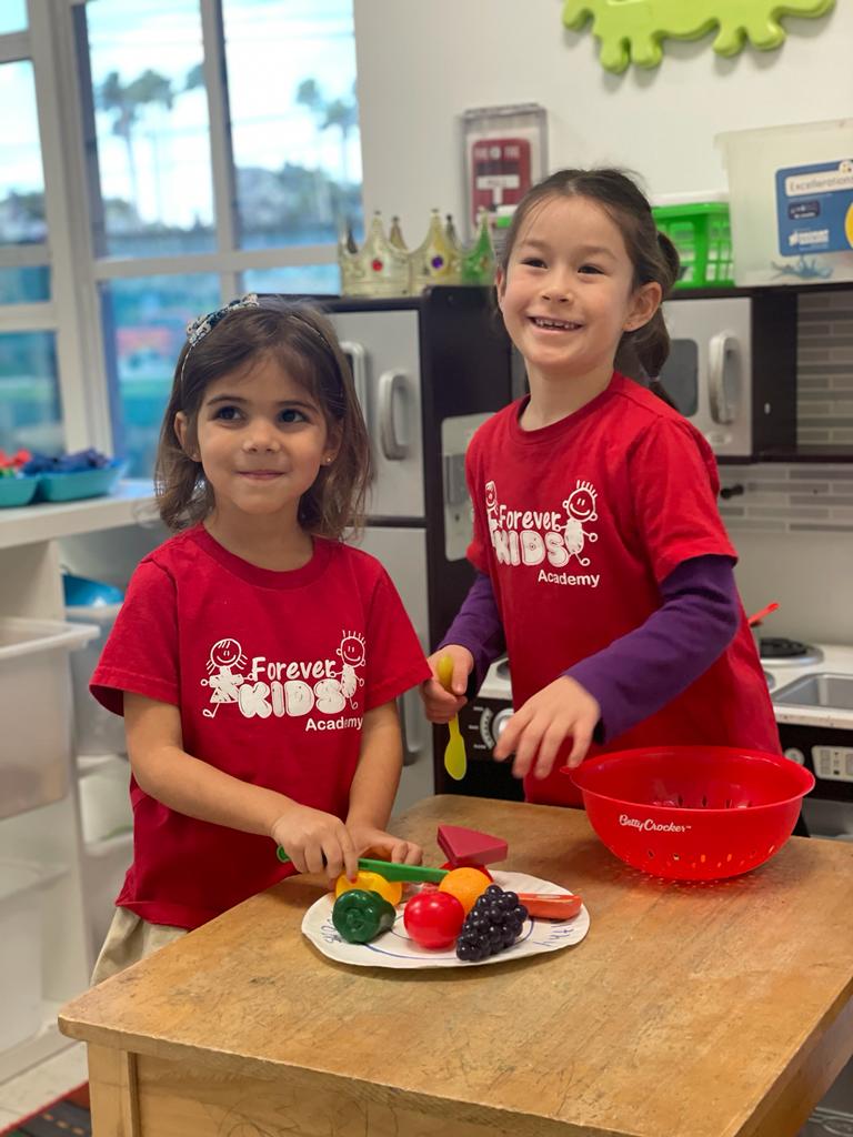 Two toddlers wearing red t-shirts with the brand Forever kids Academy text in front. Playing with play dough on a table.